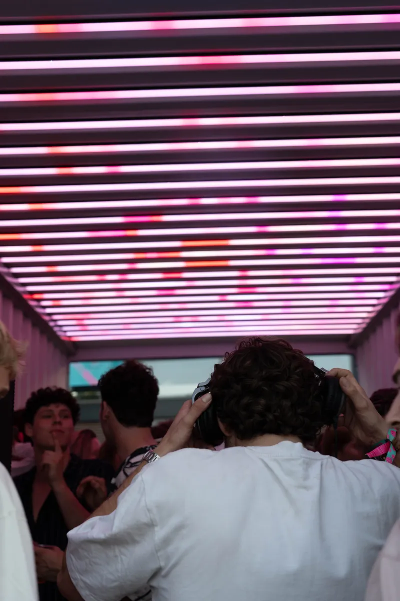DJ met koptelefoon in de RAVEBOX container onder een roze lichtshow, met feestende bezoekers in de container stage op een festival.